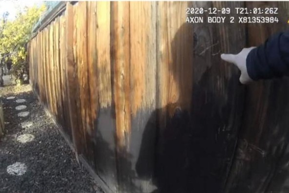 A gloved hand points to scratch marks on a wooden fence, captured on police bodycam footage from the day of the incident