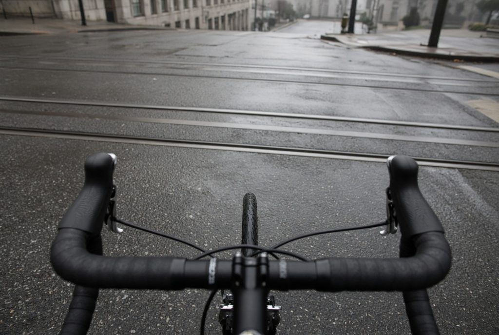 First-person view over drop handlebars as a bicycle front wheel approaches San Francisco streetcar tracks at a near 90-degree angle on wet pavement.